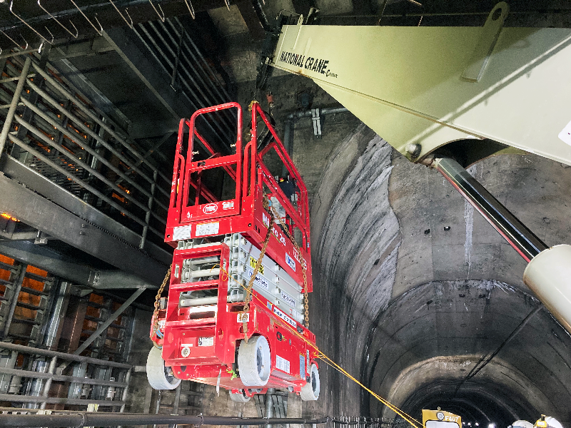 A diesel-powered work train known as a Prime Mover moves a man-lift into the trackside space prior to mesh barrier installation. This lift was used to remove the existing wall of louvers and worker platforms seen in this image.