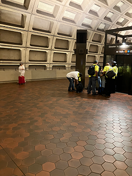 A picture of the elevator from the upper level to the lower level. Beyond the elevator, which is to the right, there is a wall. There are construction workers in front of the elevator working on the structure.
